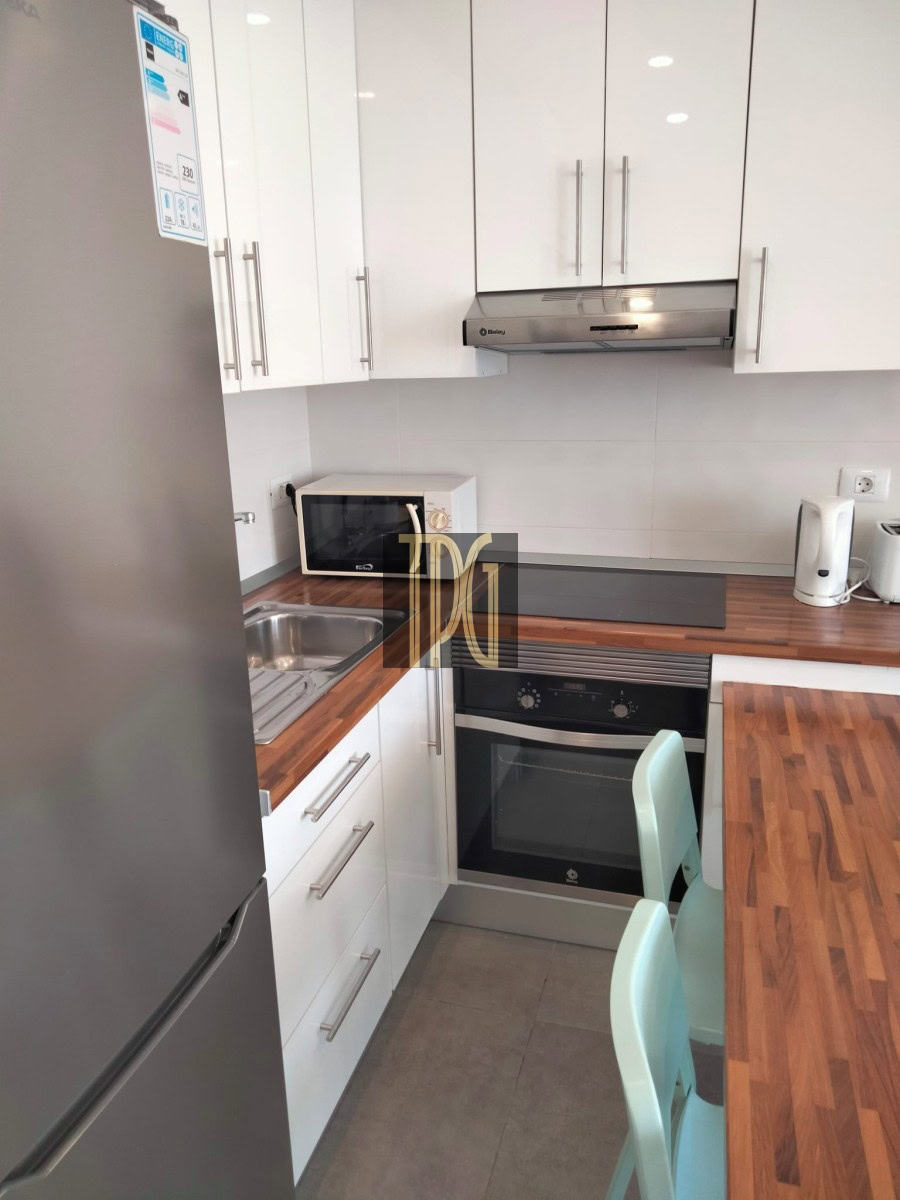 Compact L-shaped kitchen with white cabinets, stainless steel fridge, oven, and range hood on a wood countertop wall. (Informative)