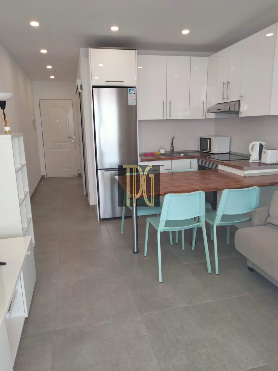 Modern open-plan kitchen with white glossy cabinets, stainless steel fridge, and a wooden island/bar area with mint teal chairs in front of a gray-tloor.