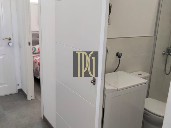 White laundry alcove between a hallway and a bathroom, with a top-loading washing machine under a water heater and a partially open door revealing a bedroom beyond.