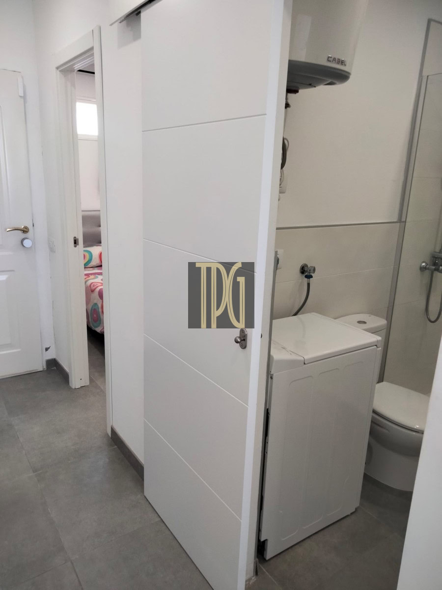 White laundry alcove between a hallway and a bathroom, with a top-loading washing machine under a water heater and a partially open door revealing a bedroom beyond.