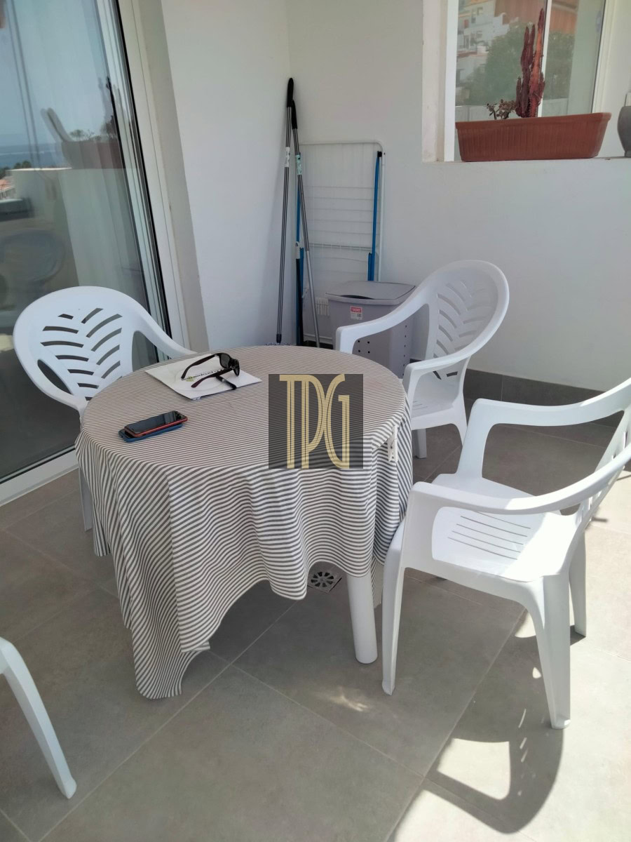 Patio balcony with a round striped table, four white plastic chairs, sunglasses, a phone on the table, and a drying rack in the background.