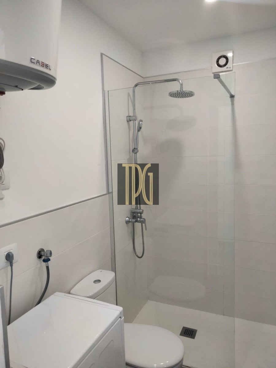 Modern bathroom with glass shower enclosure, rainfall ceiling showerhead, handheld shower, toilet, and washing machine nearby.