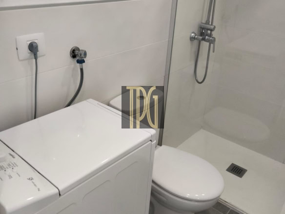 Small bathroom with a washing machine beside the toilet and a glass-enclosed shower in a white-tiled space.