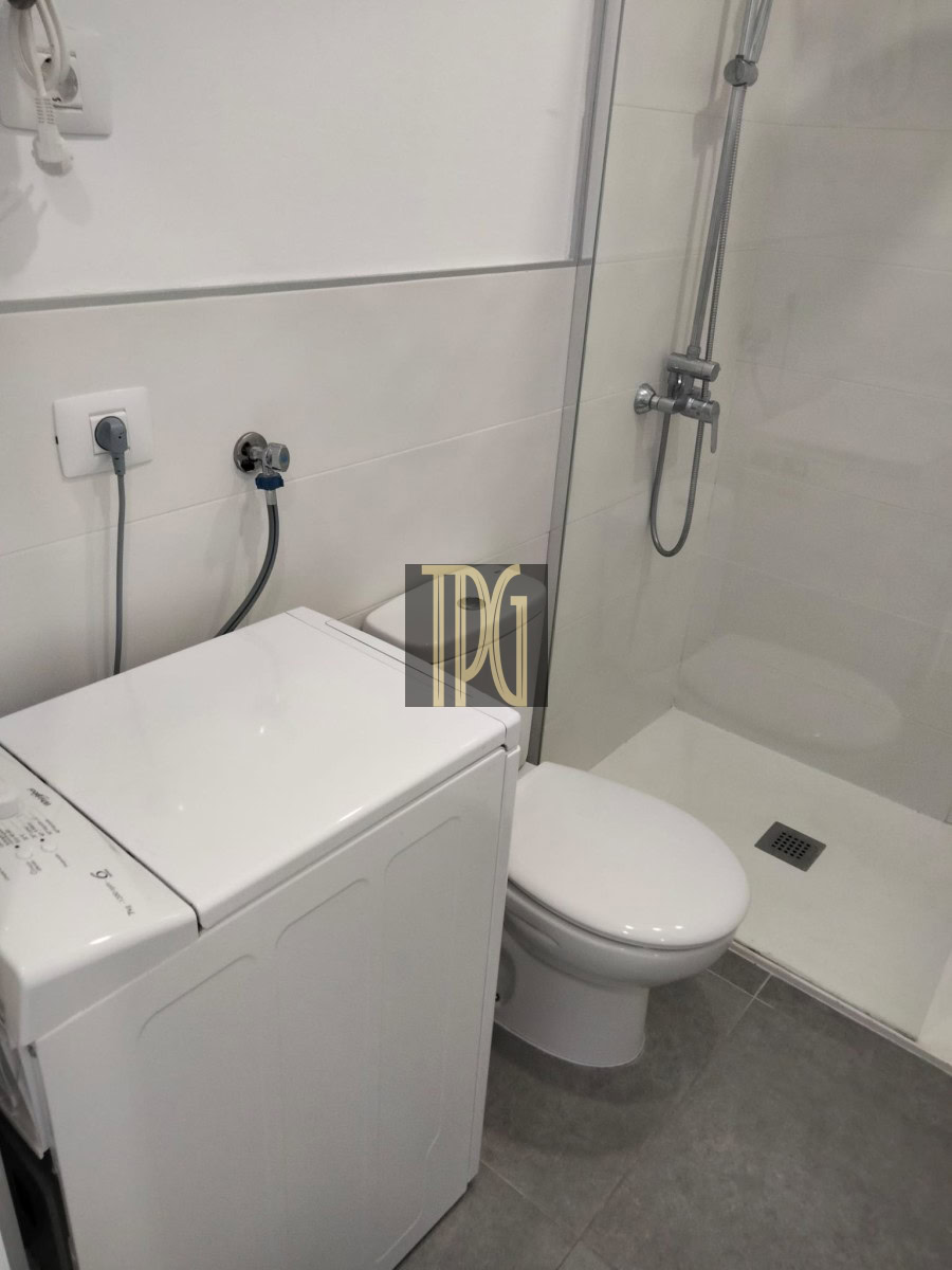 Small bathroom with a washing machine beside the toilet and a glass-enclosed shower in a white-tiled space.