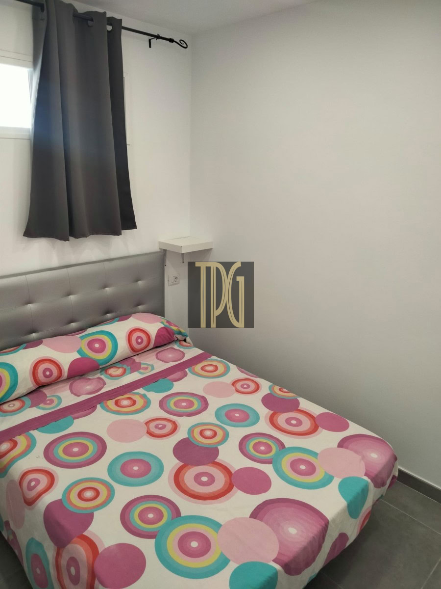 Small modern bedroom with a gray tufted headboard, white walls, and a bedspread of colorful circles (pink, teal, orange). Window with dark curtains nearby.