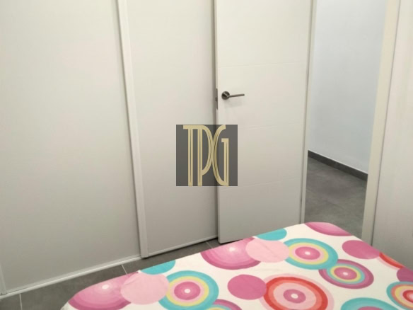 White bedroom with a closed wardrobe and an open door, bed in the foreground covered by a colorful circular-pattern duvet.