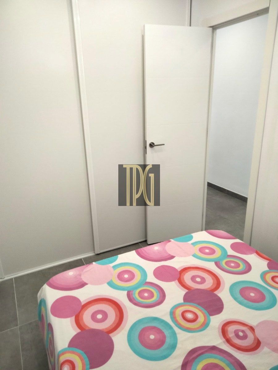 White bedroom with a closed wardrobe and an open door, bed in the foreground covered by a colorful circular-pattern duvet.
