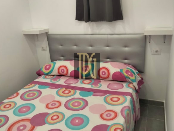 Twin bed with a tufted gray headboard and colorful circular-pattern bedding in a small white room with gray tiled floor and a window with dark curtains above the headboard.