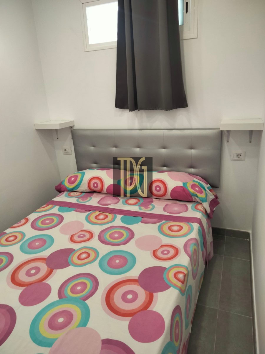 Twin bed with a tufted gray headboard and colorful circular-pattern bedding in a small white room with gray tiled floor and a window with dark curtains above the headboard.