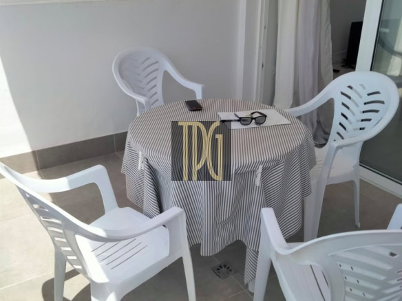 Round patio table with a striped tablecloth and four white plastic chairs on a balcony; sunglasses and a phone rest on the table.