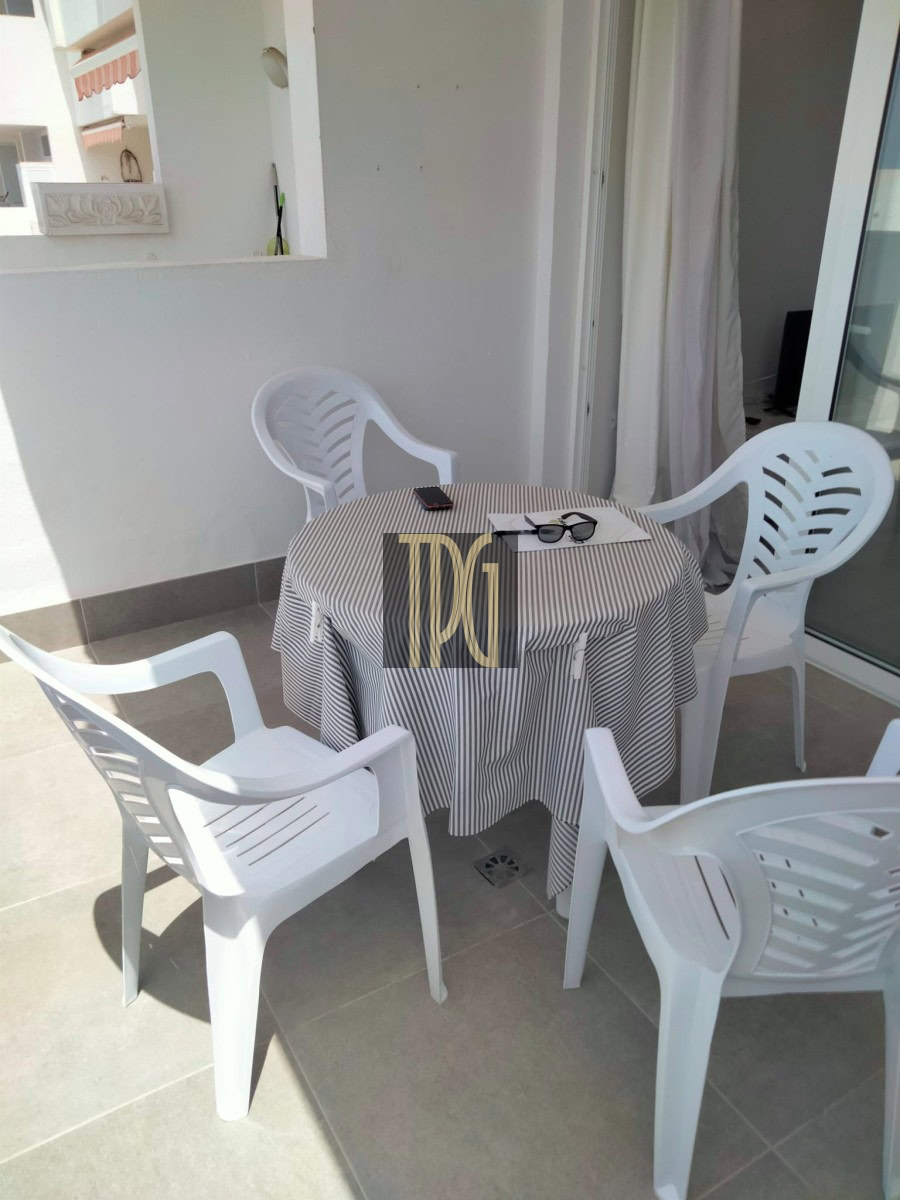 Round patio table with a striped tablecloth and four white plastic chairs on a balcony; sunglasses and a phone rest on the table.