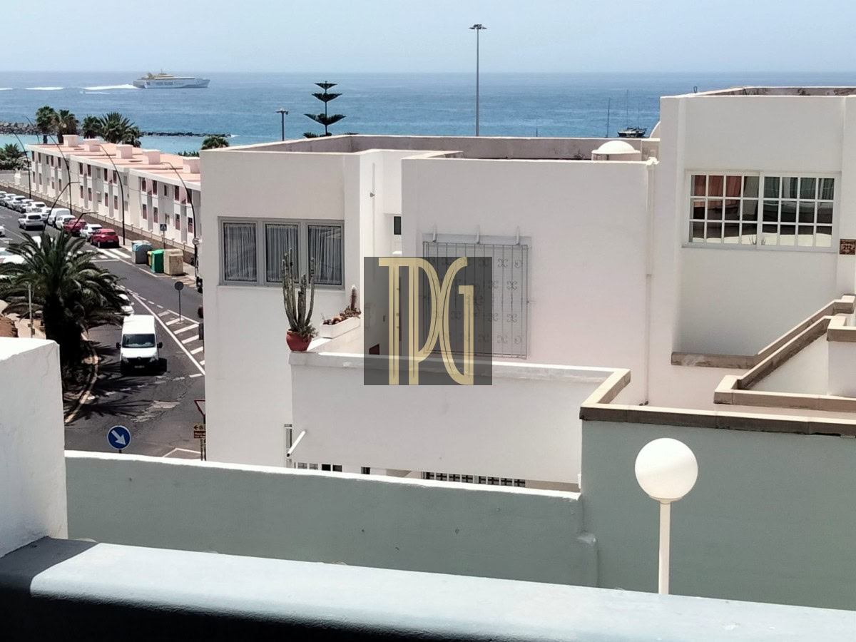White apartment buildings line a sunny coastal street with parked cars and palm trees, blue sea and a distant ferry on the horizon.