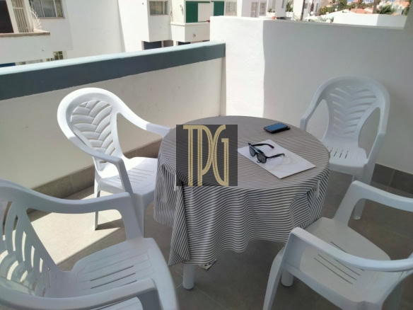 Balcony scene with four white plastic chairs around a round table covered by a striped tablecloth; a smartphone and sunglasses rest on papers.