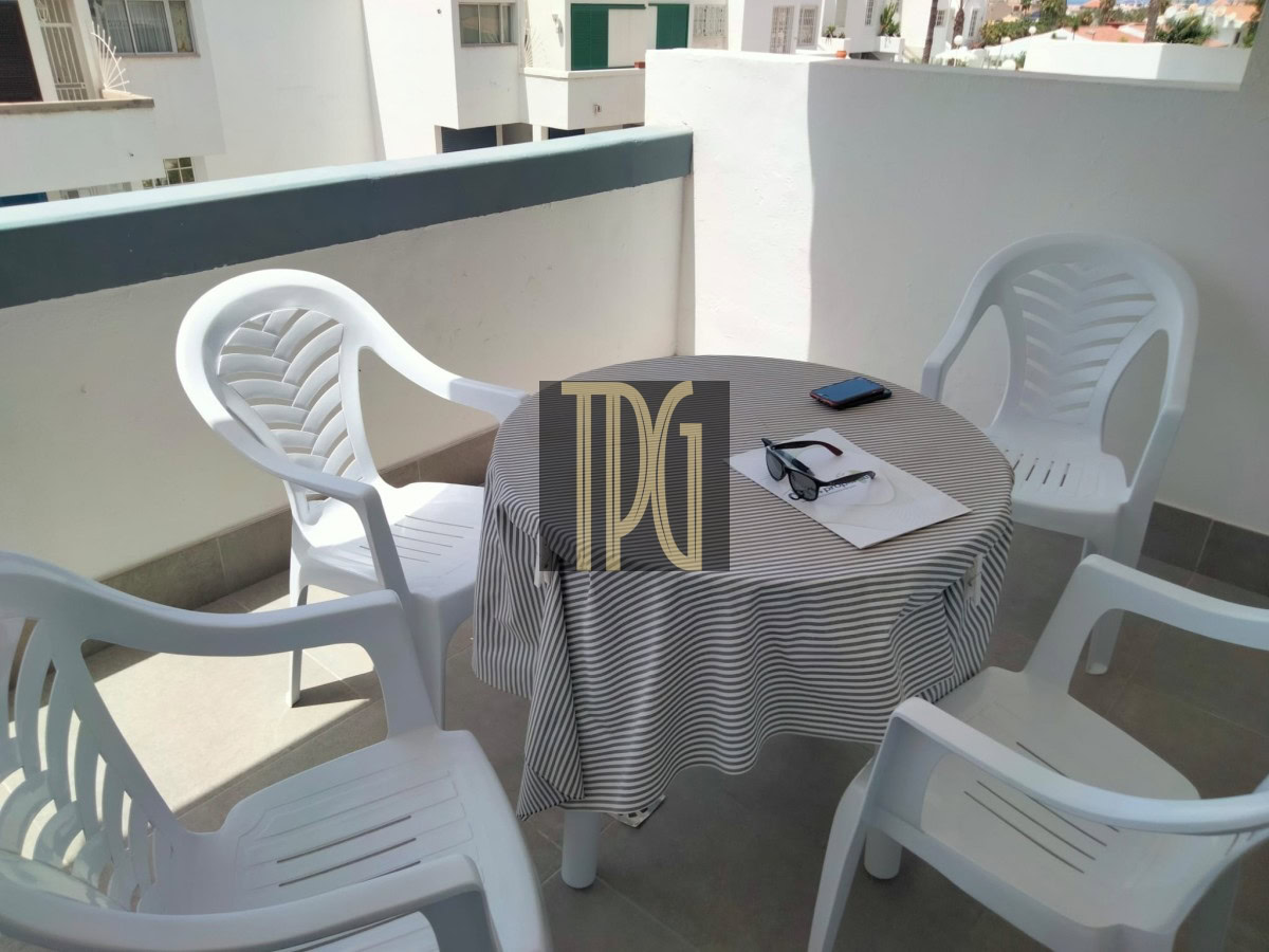 Balcony scene with four white plastic chairs around a round table covered by a striped tablecloth; a smartphone and sunglasses rest on papers.