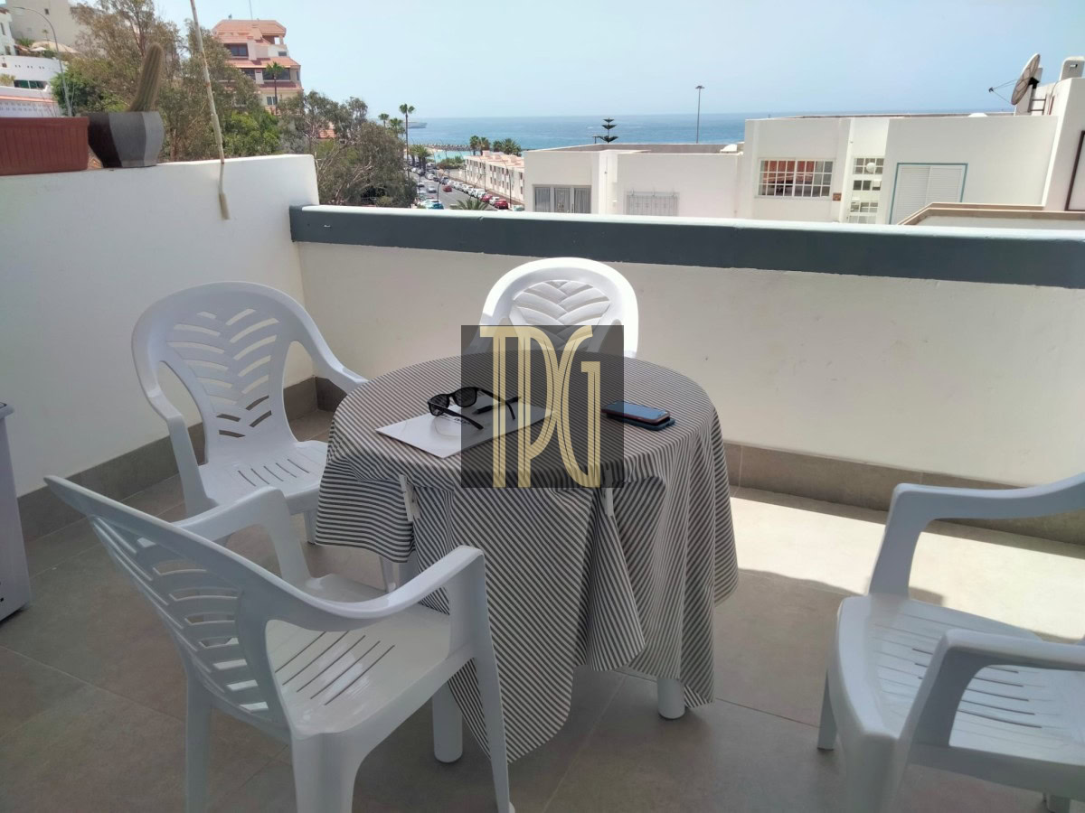 Balcony table with striped tablecloth and four white plastic chairs, ocean view in the distance, sunglasses and a phone on the table.
