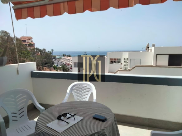 Balcony with orange striped awning, two white plastic chairs, and a round table with sunglasses and a phone, overlooking the sea.