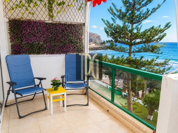 Balcony seating area with two blue lawn chairs, a small yellow table with a potted plant, and purple flower vines on a trellis, overlooking the ocean.