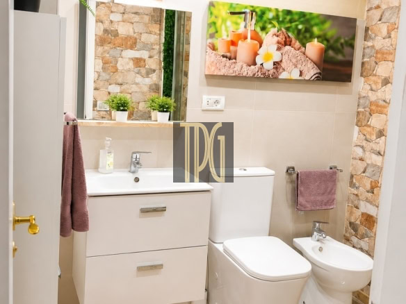 Modern bathroom with a white vanity, sink, toilet and bidet, pink rug, and stone wall accents with potted plants on shelves and warm lighting.