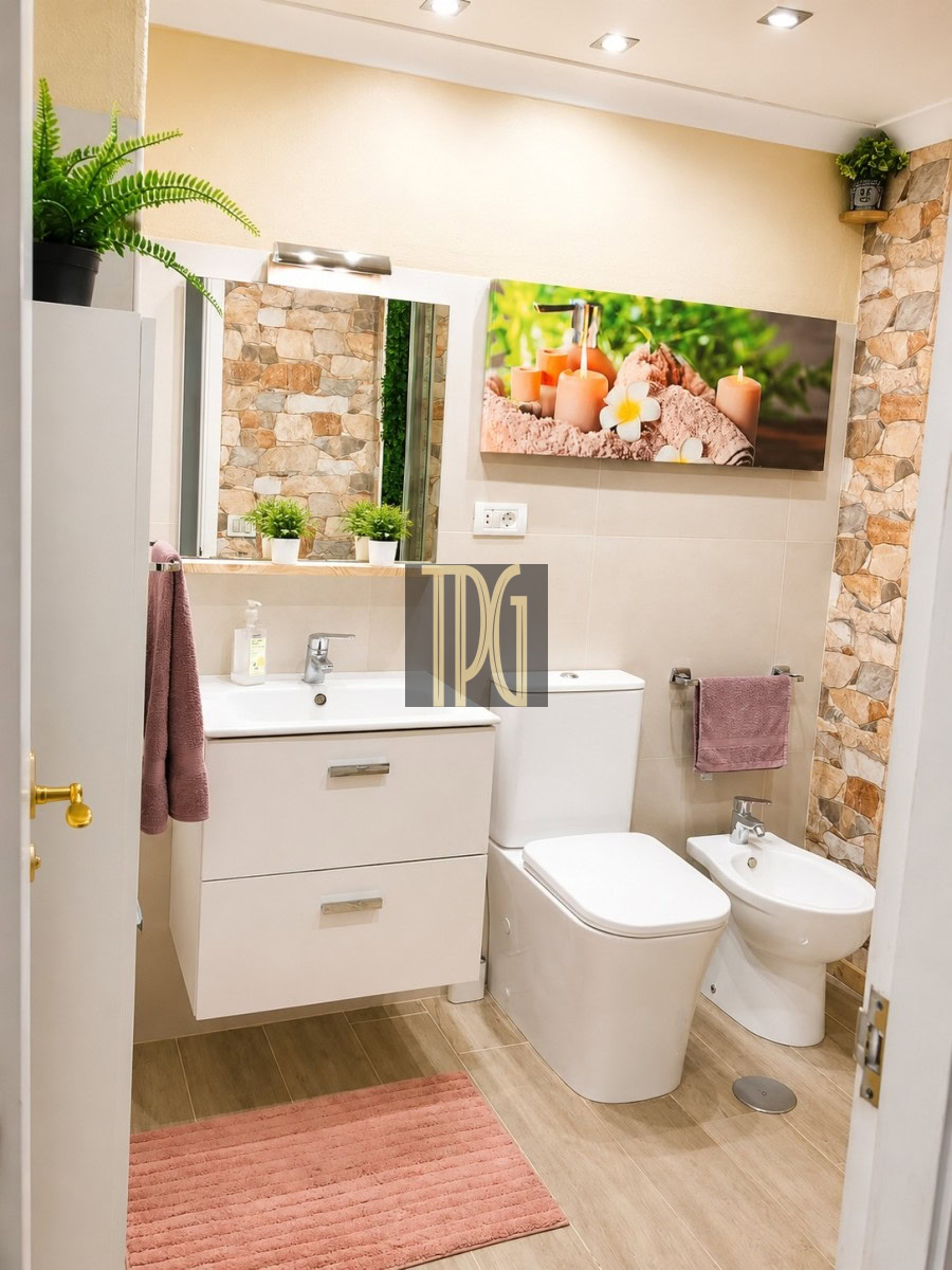 Modern bathroom with a white vanity, sink, toilet and bidet, pink rug, and stone wall accents with potted plants on shelves and warm lighting.