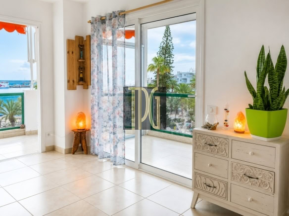 Bright living area with sliding glass balcony doors, sheer floral curtains, and a carved white dresser topped with a green plant and salt lamps.