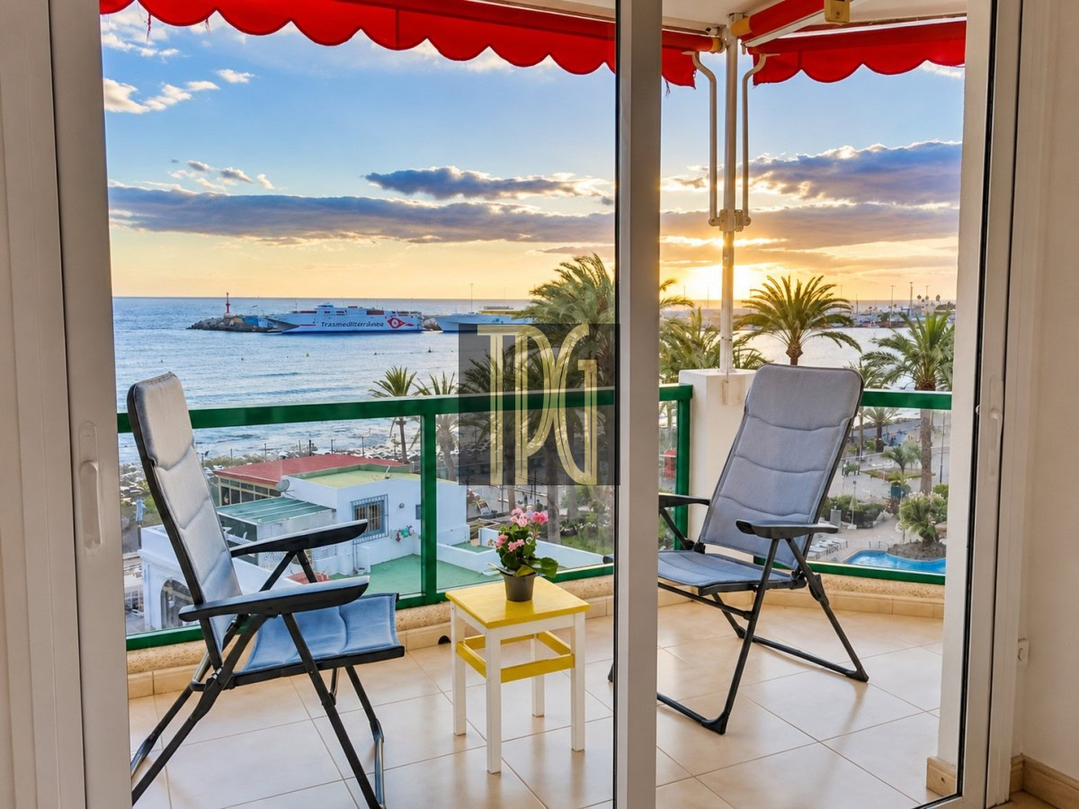 Balcony seating with two recliner chairs and a small yellow table with a potted plant, overlooking the sunset over the harbor and palm trees.
