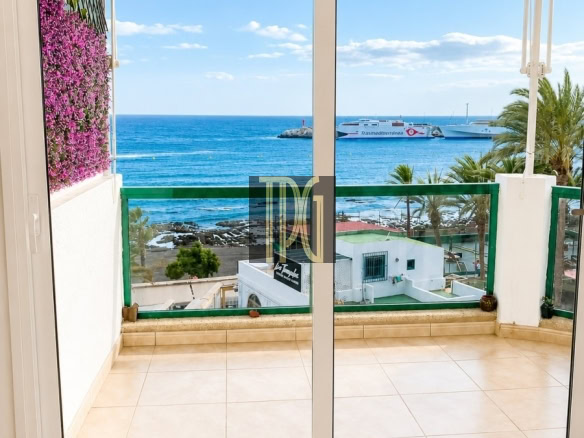View from a balcony: sliding glass doors open to a tiled terrace with a red canopy, pink flowers on the left, a green railing, and a blue sea with boats in the distance.