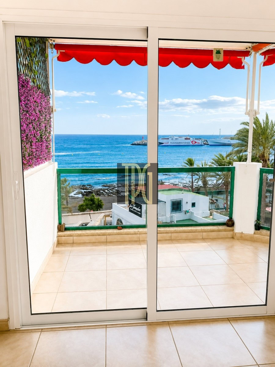 View from a balcony: sliding glass doors open to a tiled terrace with a red canopy, pink flowers on the left, a green railing, and a blue sea with boats in the distance.