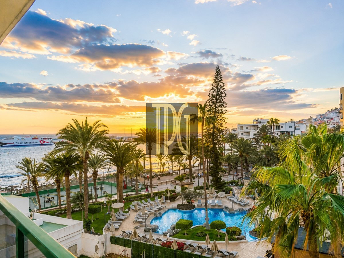 Sunset over a tropical resort with a blue swimming pool, palm trees, and lounge chairs along a garden courtyard toward the sea.