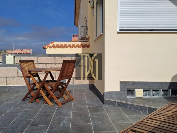 Wooden outdoor table and folding chairs on a gray tiled patio beside a beige house under a blue sky.