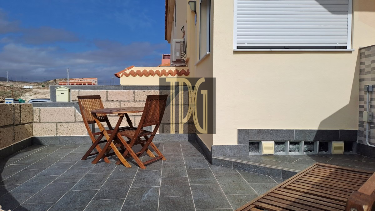 Wooden outdoor table and folding chairs on a gray tiled patio beside a beige house under a blue sky.