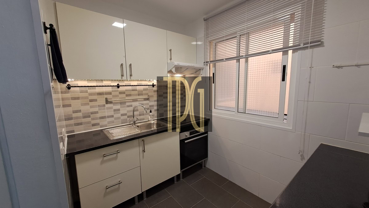 Compact kitchen with white cabinets, tiled backsplash, stainless steel sink, and dark countertop beneath a window with blinds.