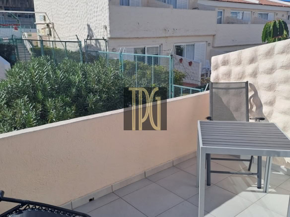 Sunlit balcony with a gray metal table and chair on tiled floor, bordered by a beige wall and lush green shrubs beyond.