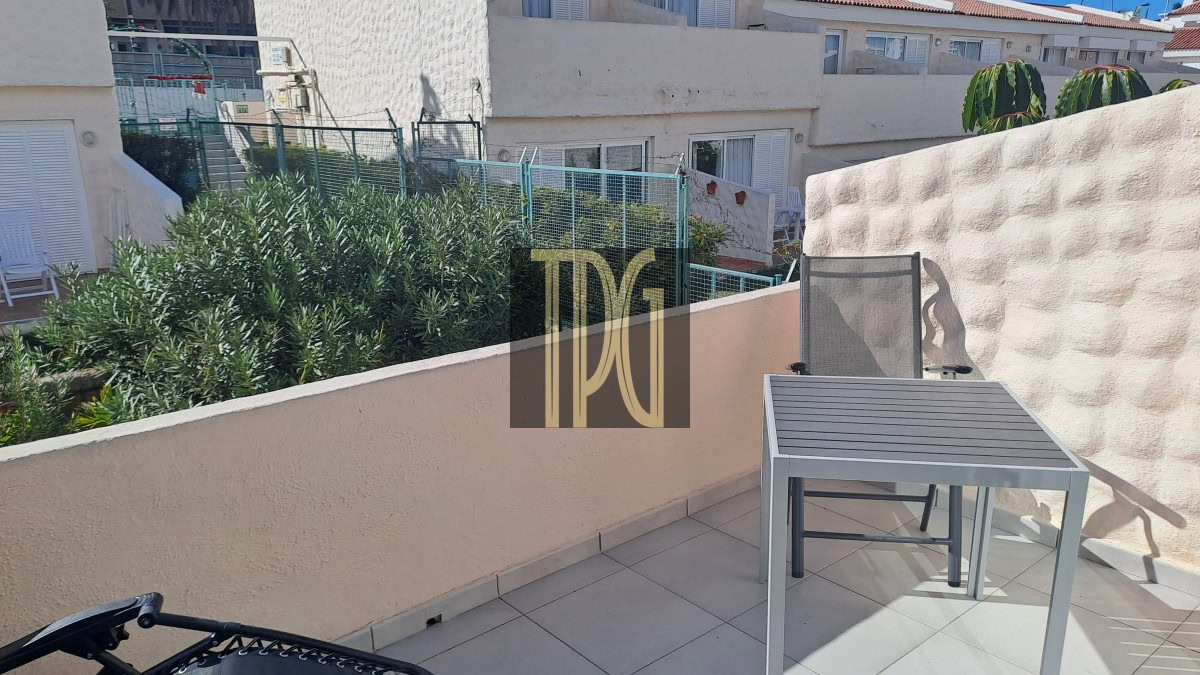 Sunlit balcony with a gray metal table and chair on tiled floor, bordered by a beige wall and lush green shrubs beyond.
