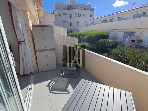 Sunlit apartment balcony with a gray storage cabinet, a metal folding chair, and a slatted table, overlooking a garden courtyard.