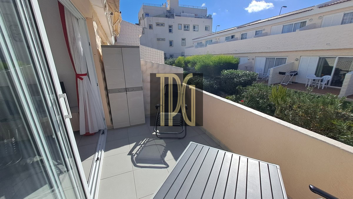 Sunlit apartment balcony with a gray storage cabinet, a metal folding chair, and a slatted table, overlooking a garden courtyard.
