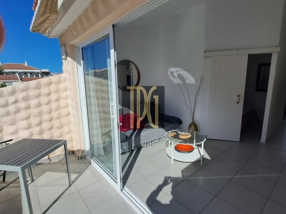 Sunny balcony with a gray table and chair, sliding glass doors open to a bright living area.