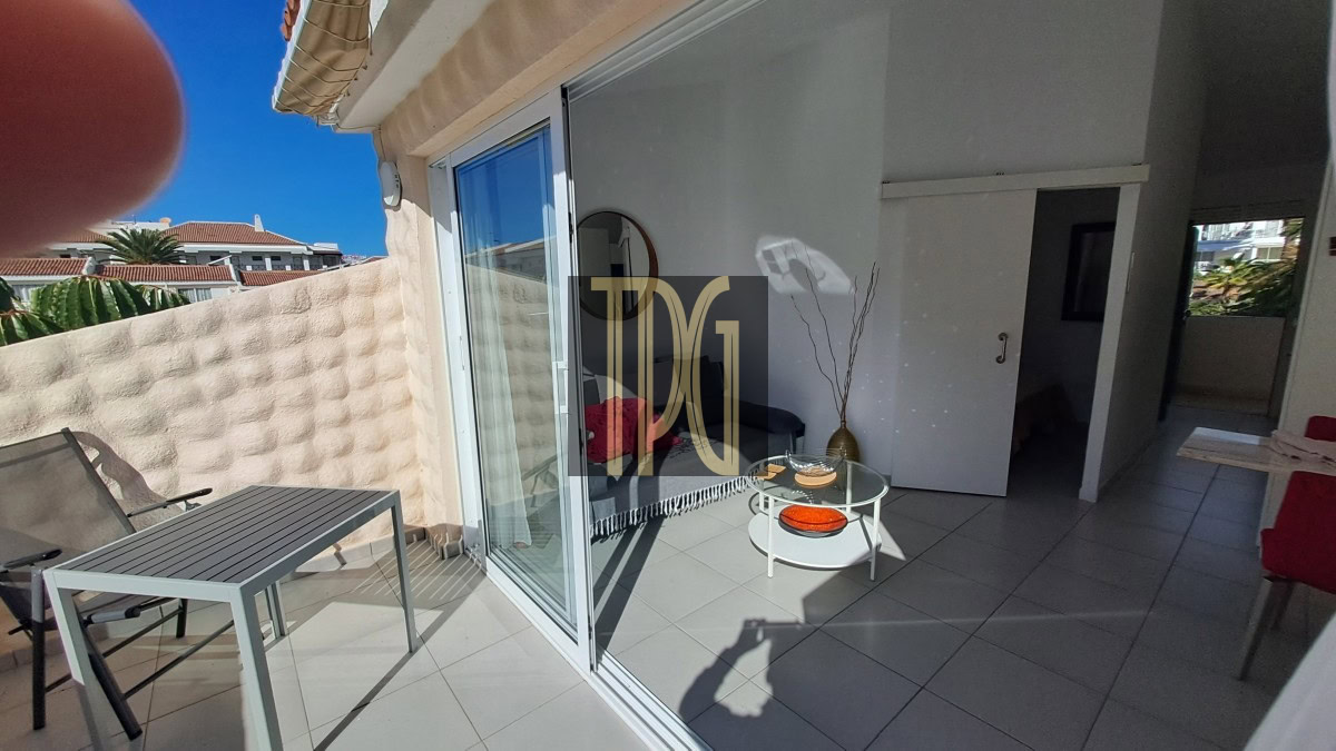 Sunny balcony with a gray table and chair, sliding glass doors open to a bright living area.