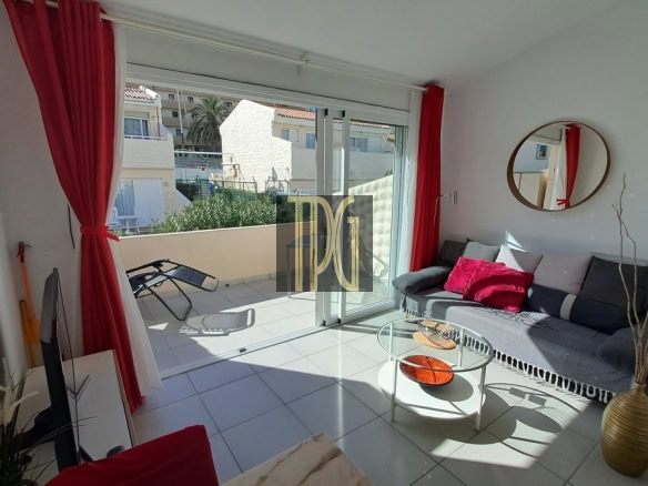 Bright living room with a gray sofa, red cushions, round wall mirror, and glass coffee table; sliding glass doors open to a sunlit balcony with a lounge chair outside.