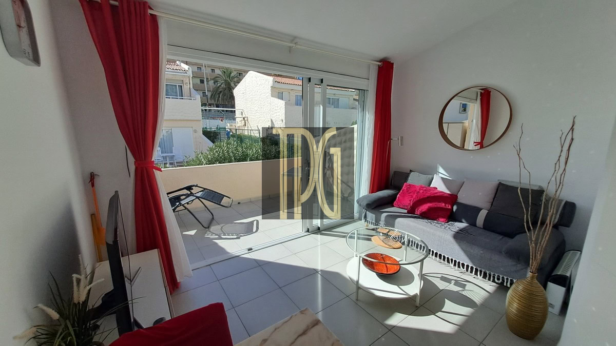 Bright living room with a gray sofa, red cushions, round wall mirror, and glass coffee table; sliding glass doors open to a sunlit balcony with a lounge chair outside.