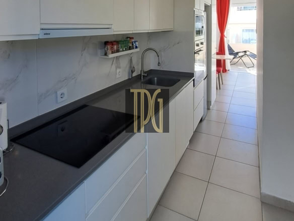 Modern white galley kitchen with gray countertop, electric cooktop, sink, and a view to a sunlit balcony with red curtains.” (informative)