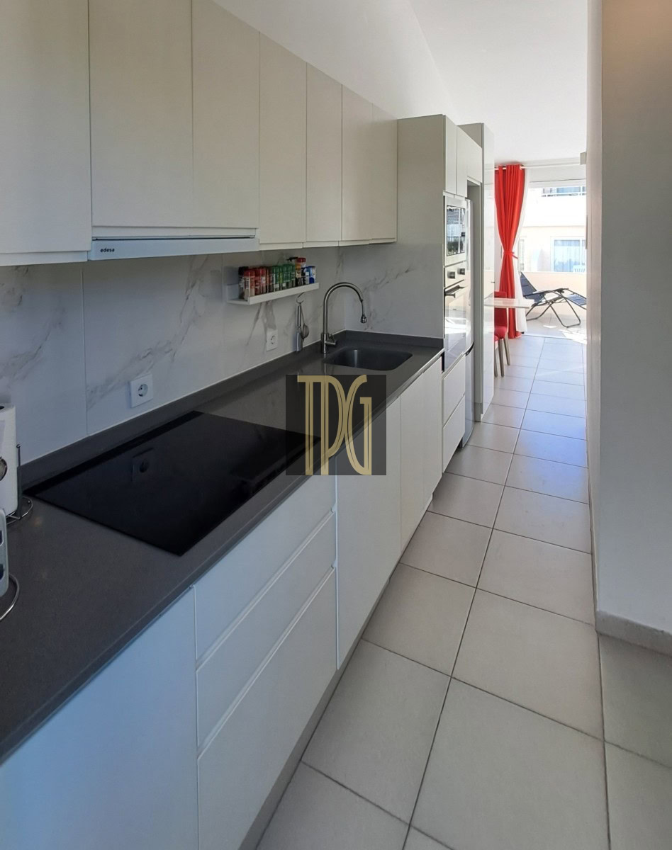 Modern white galley kitchen with gray countertop, electric cooktop, sink, and a view to a sunlit balcony with red curtains.” (informative)