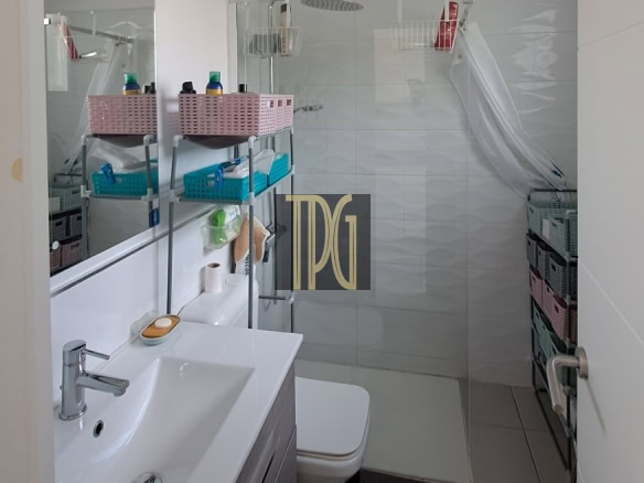 Small modern bathroom with a sink vanity, mirror, toilet, and a glass shower stall with a white curtain. Shelves hold pink baskets and blue organizers, creating organized storage.