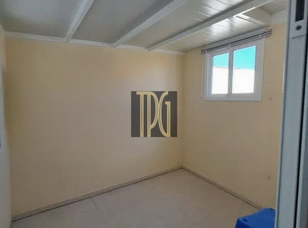 Small empty beige room with white ceiling beams, a window on the right, and a blue stool in the corner.