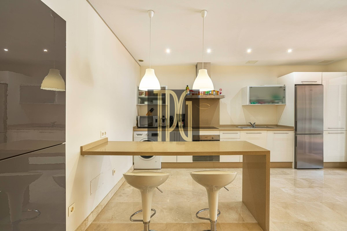 Modern open-plan kitchen with a beige island breakfast bar and two white swivel stools, stainless steel refrigerator, and built-in appliances along the back wall.