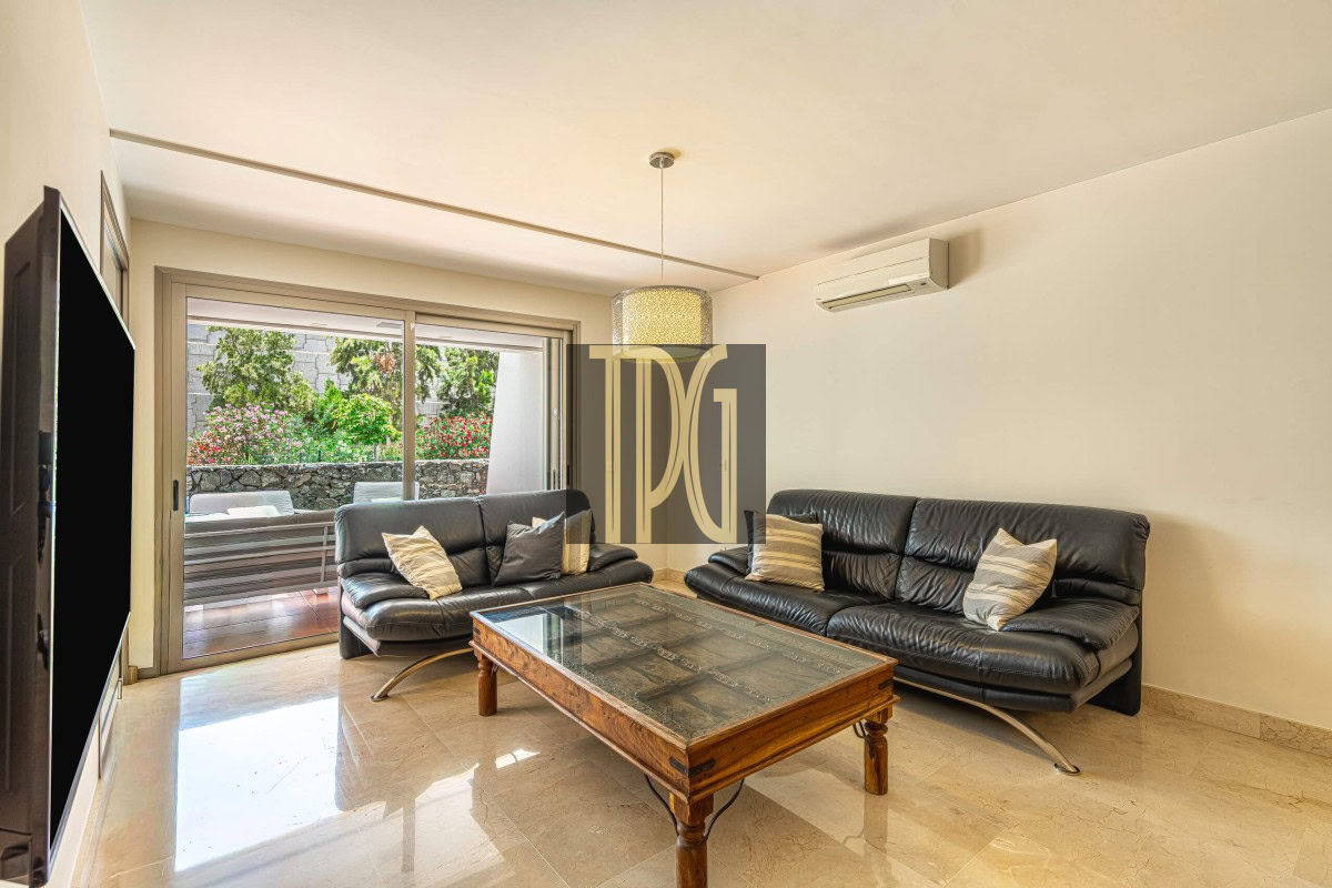 Modern living room with two black leather sofas, a glass-topped wooden coffee table, and sliding doors to a garden.