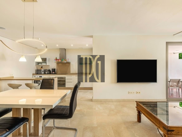 Open-plan dining area with a white table and black chairs, a modern kitchen in the background, and a flat-screen TV on the wall.