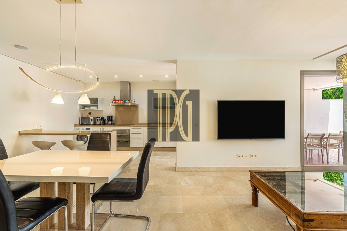 Open-plan dining area with a white table and black chairs, a modern kitchen in the background, and a flat-screen TV on the wall.