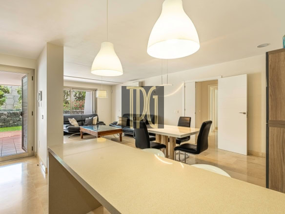 Open-plan kitchen and dining area with a beige countertop island, white walls, and black chairs; patio doors lead to a garden.