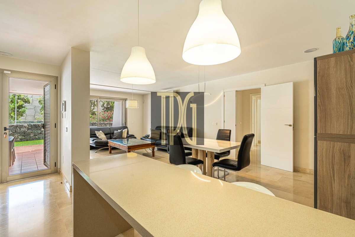 Open-plan kitchen and dining area with a beige countertop island, white walls, and black chairs; patio doors lead to a garden.