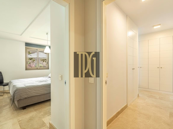 Bright hallway of a modern apartment leading to a bedroom on the left, which contains a bed, a black chair, and a window with greenery outside; white built-in closets line the right wall.